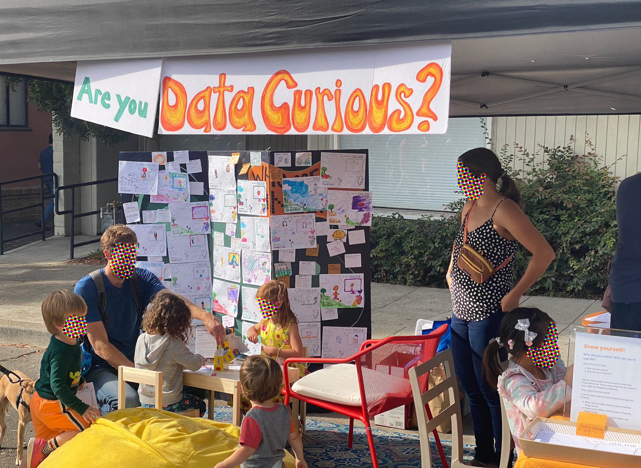 Two parents and several preschool-aged children relax in the booth. Behind them is a board covered with coloring pages made by other participants, and hanging from the sun shade at the top of the image is a sign that reads, “Are you Data Curious?”